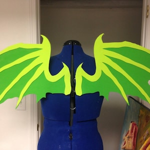 May include: A pair of green foam dragon wings with yellow accents. The wings are attached to a blue dress form.