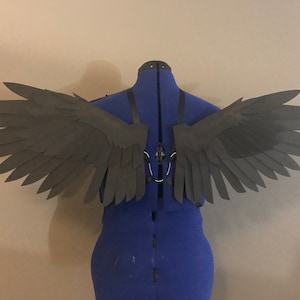 Könnte beinhalten: Dunkelgraue Kostümflügel mit geschichtetem Federdesign, ausgestellt auf einer blauen Schneiderpuppe. Die Flügel sind mit schwarzen Riemen und einem silberfarbenen Metallverschluss befestigt. Die Flügel bestehen aus einem leichten Material.