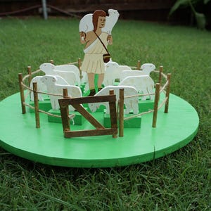 Puede incluir: Una figura de madera que representa a un pastor con una oveja sobre sus hombros, rodeado por un rebaño de ovejas dentro de una valla. La escena está ambientada sobre una base circular verde, con una puerta de madera. La figura está sobre un césped verde.