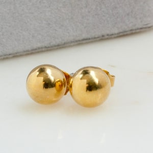 May include: A pair of gold ball stud earrings. The earrings are small and round, and they have a shiny finish.