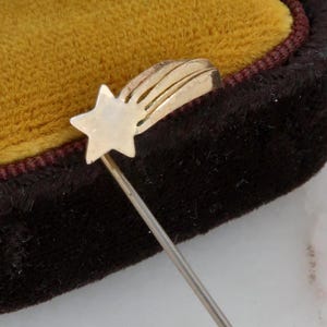 May include: A gold-toned stick pin featuring a star and comet design. The pin is displayed in a vintage brown velvet jewelry box with a mustard-colored interior. The pin's design includes a star and a comet tail.