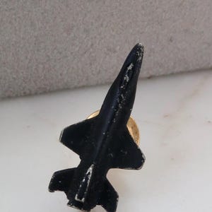 May include: A black metal airplane pin with a gold-colored pin back. The pin is shaped like a jet fighter with a detailed design. The pin is on a white surface.