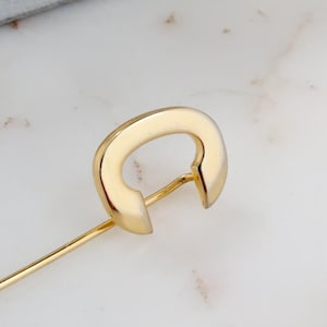 May include: A gold-colored stick pin with a rounded, open-ended design. The pin is set against a white and gray marble surface, with a gray jewelry box in the background. The pin is likely made of metal and is a jewelry item.