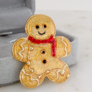 May include: A gold-coloured gingerbread man brooch with a red scarf. The brooch has a glittery finish and black button details. The gingerbread man is in a grey jewellery box.