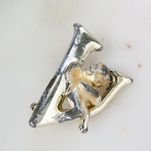 Peut inclure: Une broche dorée en forme de lettre V avec une figurine de chérubin attachée. Le chérubin est en métal blanc et a une finition dorée.
