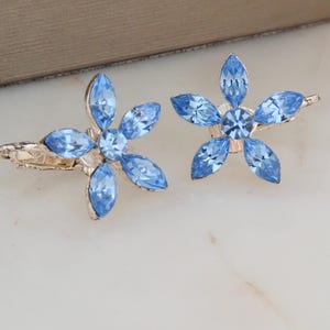May include: Two flower-shaped hair clips with light blue crystal petals and a silver-tone metal base. The floral design features five petals around a central crystal. The clips are displayed on a white surface.