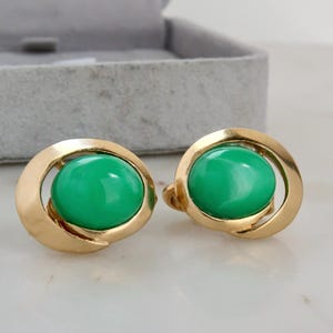 Op de afbeelding: Een paar goudkleurige oorbellen met een heldergroene, ovaalvormige middensteen. De oorbellen hebben een cirkelvormig ontwerp met een gebogen, open frame. De oorbellen worden gepresenteerd op een wit oppervlak.