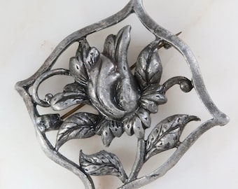 May include: A silver-toned brooch featuring a detailed floral design with a single flower and leaves. The flower is in the center of the brooch and is surrounded by a delicate, openwork frame.