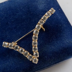 Peut inclure: Une broche dorée en forme de V, sertie de strass transparents. La broche est présentée sur un fond de velours bleu.