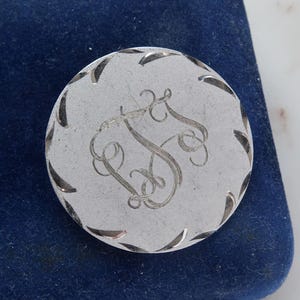 Puede incluir: Un objeto redondo plateado con un borde decorativo e iniciales grabadas. El objeto está sobre una superficie azul oscuro, similar al terciopelo. Las iniciales parecen ser "RS".