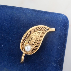 May include: A gold leaf brooch with a pearl in the center, displayed on a blue velvet background.