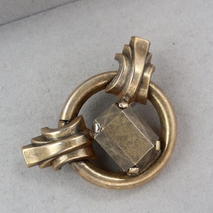 May include: A vintage-style brooch in a gold tone, featuring a geometric design. The brooch has a circular element, a square-shaped center, and decorative, curved accents. The piece is set against a neutral gray background.