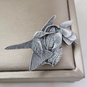以下が含まれることがあります： 羽根が細かく表現された銀色のハチドリのブローチ。花柄のデザインが施されています。ハチドリは花から蜜を吸っているように、くちばしが伸びた状態で飛んでいる様子が描かれています。