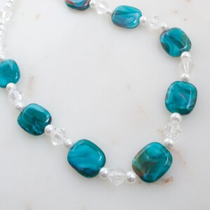 May include: A necklace featuring turquoise and brown glass beads interspersed with clear crystal and small white pearl beads. The necklace has a delicate appearance and is strung on a white cord.