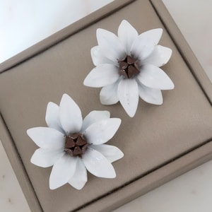 Puede incluir: Un par de pendientes de flores de esmalte blanco con centros marrones, expuestos en una caja de joyería de terciopelo gris.