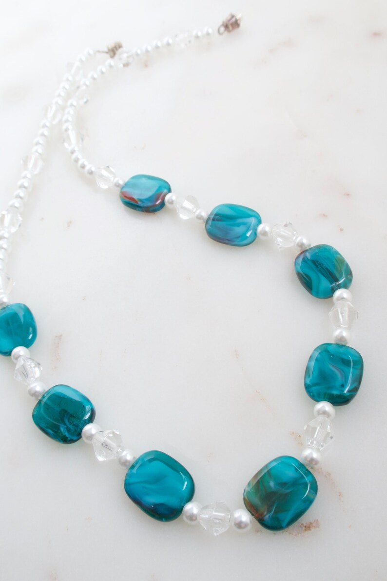 May include: A necklace featuring turquoise and brown glass beads interspersed with clear crystal and small white pearl beads. The necklace has a delicate appearance and is strung on a white cord.