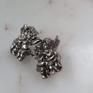 K&ouml;nnte beinhalten: Zwei silberfarbene Cherubim-Figuren. Jeder Cherub hat detaillierte Fl&uuml;gel und befindet sich in einer Umarmungspose. Die Figuren befinden sich auf einer wei&szlig;en Oberfl&auml;che. Die Cherubim sind klein, detailliert und haben eine Vintage-&Auml;sthetik.