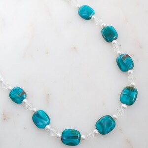 May include: A necklace featuring turquoise and clear beads. The necklace has a delicate design, with rectangular turquoise beads interspersed with small clear beads and tiny white pearls. The beads are strung on a thin, white cord.