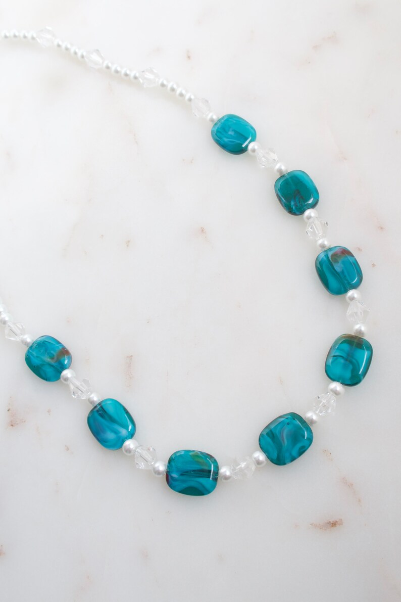 May include: A necklace featuring turquoise and clear beads. The necklace has a delicate design, with rectangular turquoise beads interspersed with small clear beads and tiny white pearls. The beads are strung on a thin, white cord.