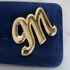Może przedstawiać: Złota metalowa broszka w kształcie litery "M" spoczywa na ciemnoniebieskiej aksamitnej powierzchni. Broszka ma błyszczące wykończenie i kursywny design, sugerujący styl vintage lub klasyczny. Tło to marmurowa powierzchnia.