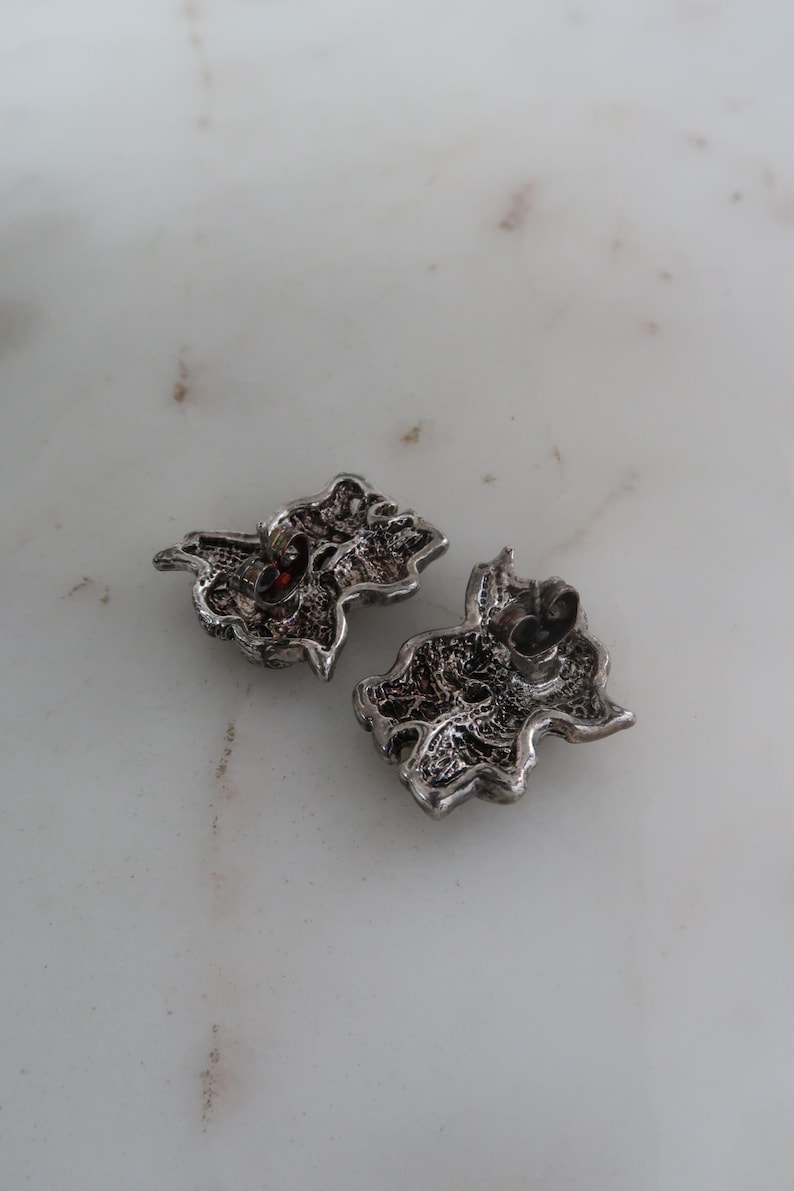K&ouml;nnte beinhalten: Zwei silberfarbene, verzierte Ohrstecker. Jeder Ohrring hat ein detailliertes, strukturiertes Design mit einem kleinen, roten Akzent. Die Ohrringe sind wie stilisierte Bl&auml;tter oder Blumenmotive geformt, mit einem Stift zum Befestigen.