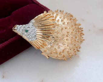 May include: A gold-toned brooch in the shape of a hedgehog with a green eye and clear rhinestones. The hedgehog's body is textured and the spines are made of gold-toned metal.