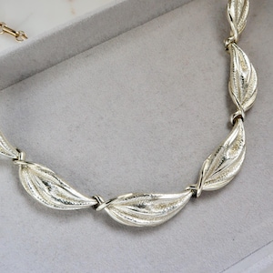 Puede incluir: Un collar de plata con un diseño de hojas. El collar está hecho de hojas unidas que están retorcidas.