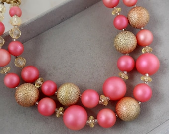 May include: A two-strand necklace featuring pink and gold beads. The necklace has large, round, matte pink beads interspersed with textured gold beads and smaller clear beads. The necklace is displayed on a gray surface.