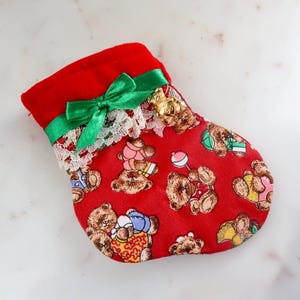 May include: A red Christmas stocking with a green bow and white lace trim. The stocking is decorated with a pattern of teddy bears wearing different outfits. There is a gold bell charm attached to the stocking.