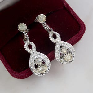 Op de afbeelding: Zilverkleurige oorhangers met een textuur infinity symbool ontwerp. Elke oorbel heeft een kleine, ronde, heldere edelsteen aan de bovenkant en drie in de infinity lus. De oorbellen worden getoond tegen een bordeauxrode fluwelen achtergrond.