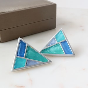 May include: A pair of geometric earrings with a silver-tone frame. Each earring features a triangular shape with sections of blue, teal, and light blue enamel. A brown box is in the background.