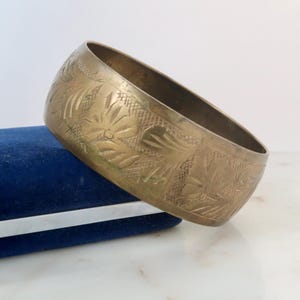May include: A wide, brass-colored bangle bracelet with an etched floral design. The bracelet is resting on a blue velvet surface. The bracelet's surface is detailed with a repeating pattern of leaves and flowers.
