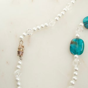 May include: A delicate necklace featuring white faux pearls, clear crystal beads, and square turquoise beads. The necklace has a magnetic clasp and is displayed on a white marble surface. The jewelry is a classic accessory.