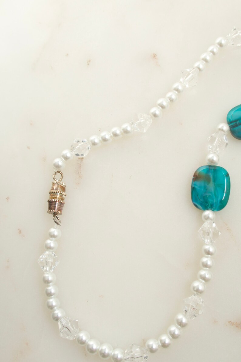 May include: A delicate necklace featuring white faux pearls, clear crystal beads, and square turquoise beads. The necklace has a magnetic clasp and is displayed on a white marble surface. The jewelry is a classic accessory.