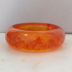 May include: A chunky, orange-colored bangle bracelet. The translucent resin has a marbled effect with swirls of darker orange and brown. The bracelet is a bold, statement piece.