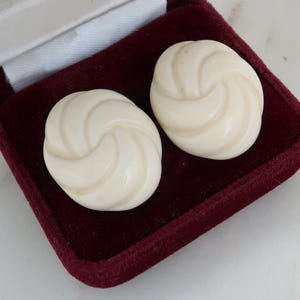 Può includere: Un paio di orecchini color crema con un design a spirale. Gli orecchini rotondi sono in un portagioie di velluto bordeaux. Gli orecchini hanno una finitura lucida. Il portagioie è aperto, rivelando gli orecchini.