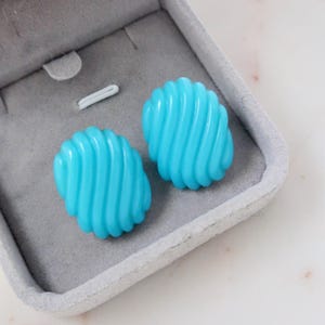 Puede incluir: Dos pendientes turquesa cuadrados con un diseño en espiral, presentados en una caja de joyería gris. Los pendientes tienen un acabado brillante y una superficie texturizada. La caja de joyería está abierta, revelando los pendientes.