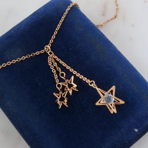 May include: A gold-colored necklace with a chain and star pendants. The necklace features three small star charms and a larger star pendant with a clear, faceted stone center. The necklace is displayed on a dark blue velvet surface.