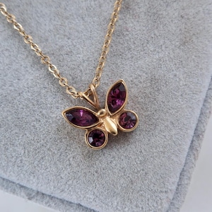 Può includere: Una collana color oro con un ciondolo a forma di farfalla. Il ciondolo è decorato con quattro pietre sfaccettate color viola intenso disposte a formare le ali, con un corpo in metallo color oro liscio al centro, appeso a una catena.