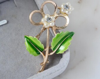 May include: A gold-tone flower brooch with white pearl-like petals and a central rhinestone. The brooch features green enamel leaves and a stem with thorns. The brooch is displayed in a gray jewelry box.