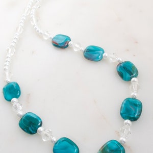 May include: A necklace featuring large, teal-colored, rectangular beads with swirling patterns. The necklace also includes smaller clear crystal and white pearl beads, strung together with a delicate chain. The necklace is a jewelry item.