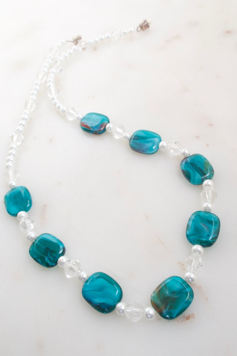 May include: A necklace featuring large, teal-colored, rectangular beads with swirling patterns. The necklace also includes smaller clear crystal and white pearl beads, strung together with a delicate chain. The necklace is a jewelry item.