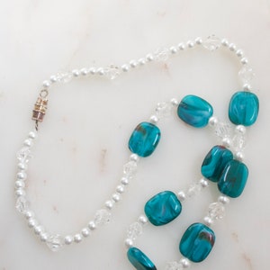 May include: A necklace featuring alternating white faux pearls, clear crystal beads, and rectangular teal glass beads. The necklace has a silver-toned magnetic clasp. The beads are arranged in a repeating pattern.