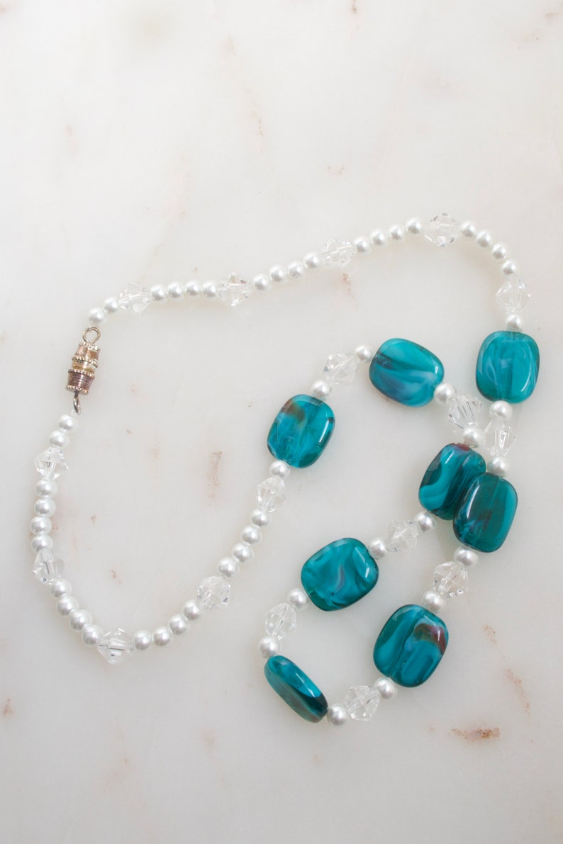 May include: A necklace featuring alternating white faux pearls, clear crystal beads, and rectangular teal glass beads. The necklace has a silver-toned magnetic clasp. The beads are arranged in a repeating pattern.