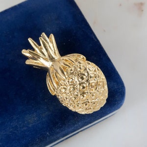 Op de afbeelding: Een gouden ananas broche met een getextureerd oppervlak. De broche ligt op een blauwe fluwelen achtergrond.