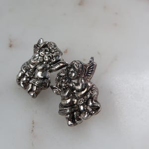 K&ouml;nnte beinhalten: Zwei silberfarbene Cherubim-Figuren. Jeder Cherub hat detaillierte Fl&uuml;gel und befindet sich in einer Umarmungspose. Die Figuren befinden sich auf einer wei&szlig;-grauen Marmoroberfl&auml;che.