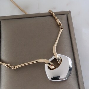 May include: A gold-toned necklace with a silver-colored square pendant. The pendant is connected to the chain by two gold-toned curved bars. The necklace is displayed on a brown surface.