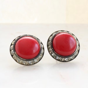 May include: A pair of red and silver stud earrings. The earrings are round and have a silver rim with small clear rhinestones around the edge. The center of the earrings are red.