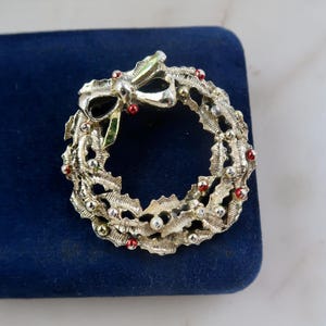 May include: Silver-toned Christmas wreath brooch with holly leaves, red berry accents, and a bow. The brooch is set on a dark blue velvet surface. A festive holiday accessory.