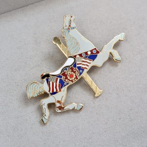 May include: A white carousel horse brooch with red, white, and blue accents. The horse is standing on its hind legs and has a gold pole attached to its back.